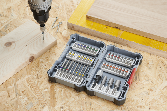 Drill driving a screw into wood beside an open case of assorted bits.