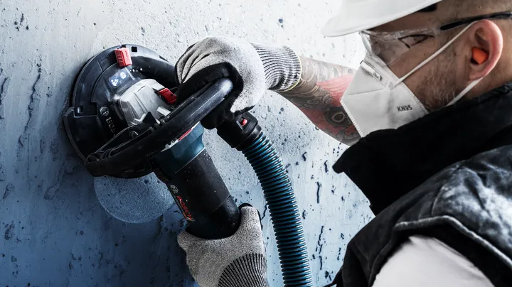 A person wearing safety equipment sands a concrete wall using a power tool.