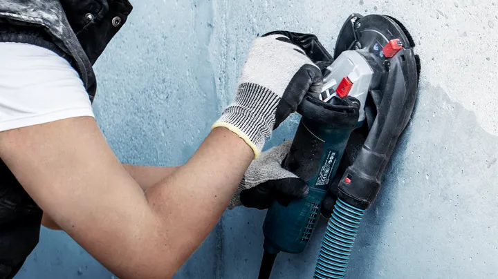 Person wearing safety equipment sands a wall with a power tool.