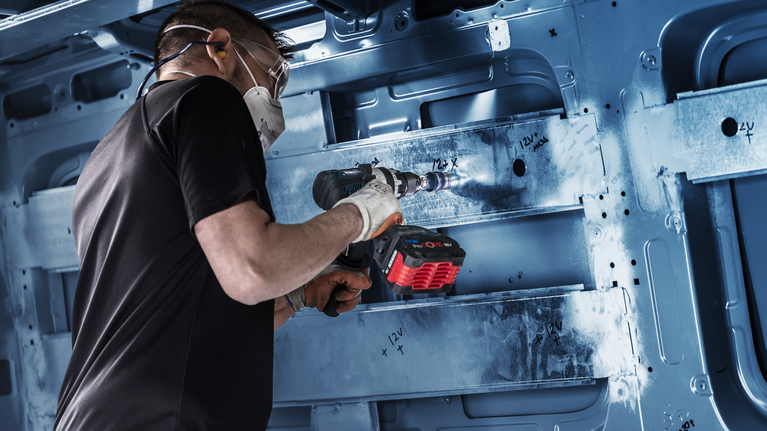 A person wearing safety equipment drills a circular hole into a metal frame.