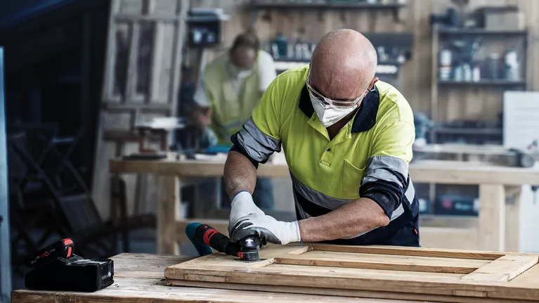 A person wearing safety equipment sands a wooden frame with a power tool.