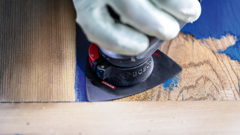 A person wearing safety equipment sands paint from wood using a power tool.