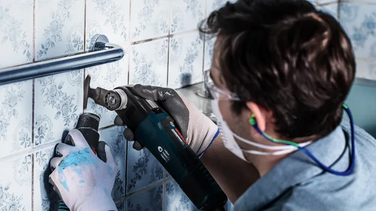 Person wearing safety equipment cuts tile grout with a power tool.