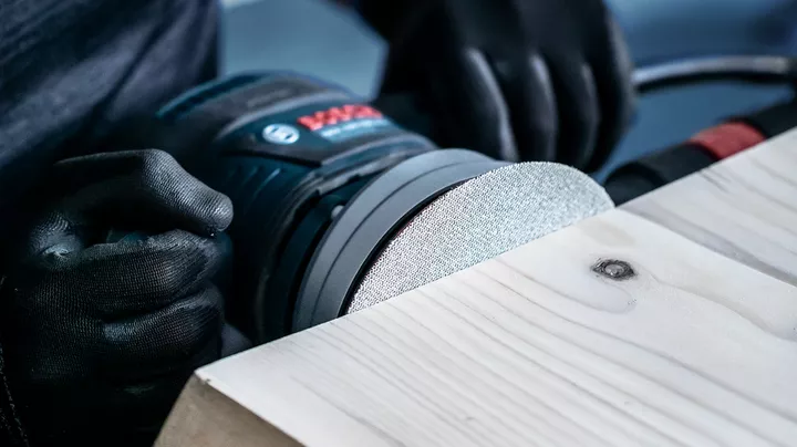 Person wearing safety equipment sands a wooden surface with a power tool.