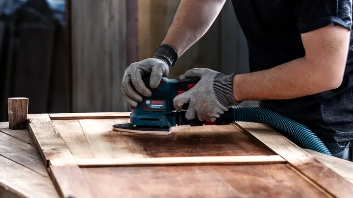 Person wearing safety equipment sanding a wooden door with a power tool.