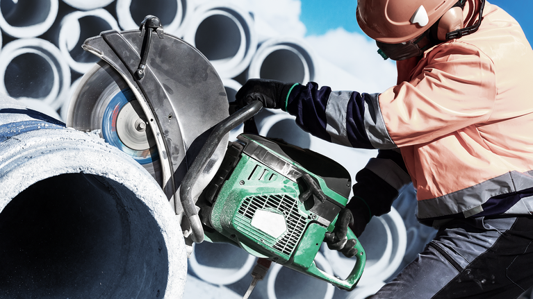 A person wearing safety equipment cuts a concrete pipe with a large power saw.