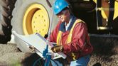 A person wearing safety equipment uses a measuring wheel while inspecting blueprints.