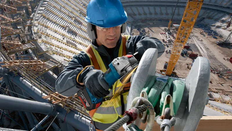 A worker wearing safety equipment uses a cordless angle grinder on a metal beam.