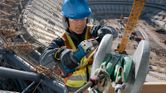 A worker wearing safety equipment uses a cordless angle grinder on a metal beam.