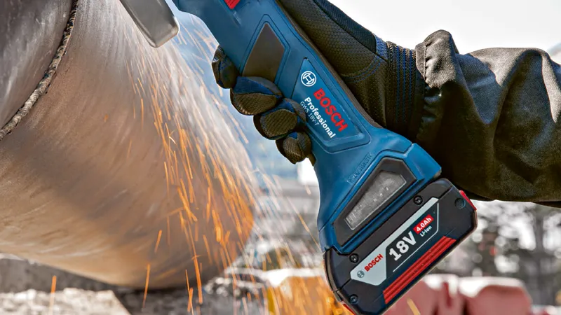 A person wearing safety equipment grinds a metal pipe with a cordless angle grinder.
