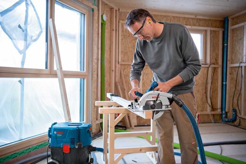 A person wearing safety equipment cuts wood with a cordless circular saw indoors.