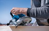 A person guides a cordless circular saw along a wooden plank.