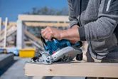 A person cuts a wooden beam outdoors using a cordless circular saw.