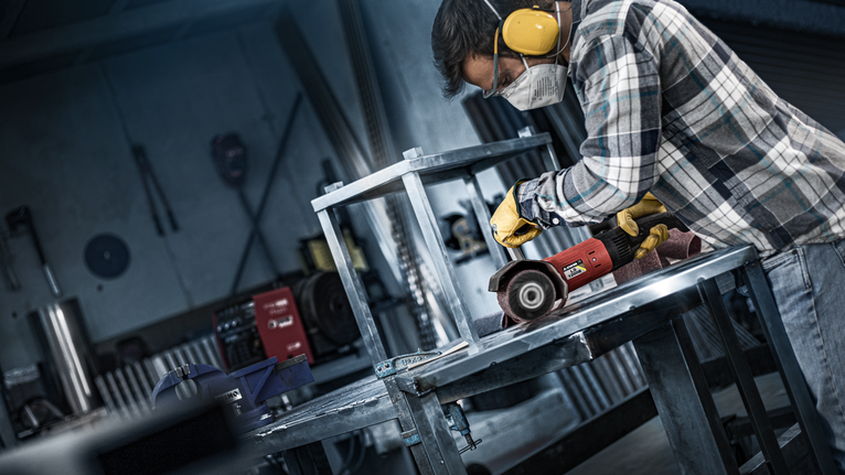 A person wearing safety equipment grinds metal on a workbench.