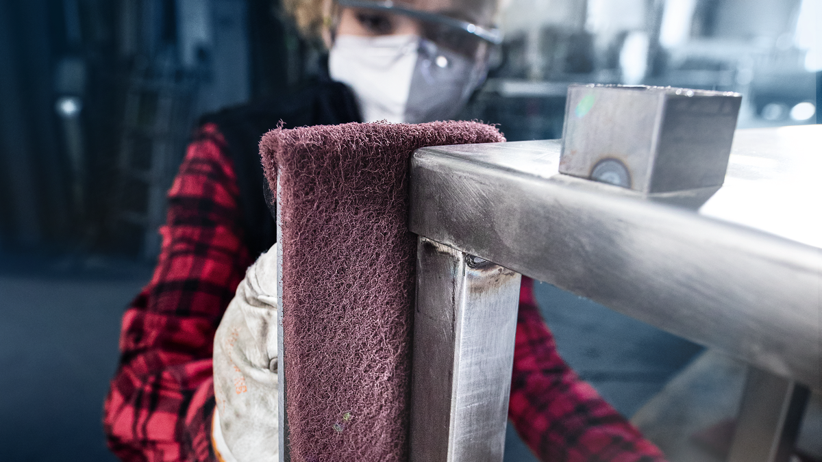 Person wearing safety equipment polishes a metal frame with a sanding tool.
