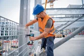 A person wearing safety equipment uses a cordless impact wrench to secure a metal fence.