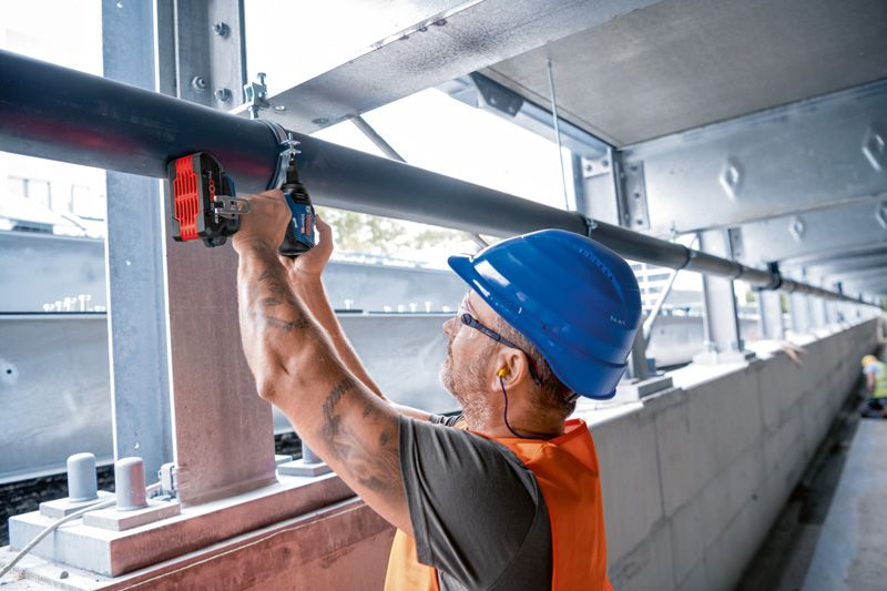 A person wearing safety equipment uses an impact wrench to fasten a metal pipe.