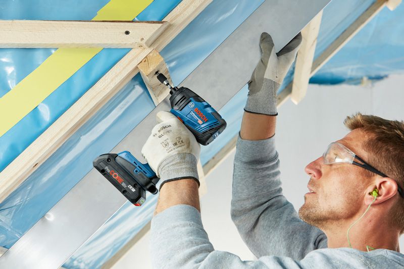 A person wearing safety equipment uses a cordless drill to fasten a metal beam to wood.