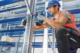 A person wearing safety equipment uses a cordless drill to assemble metal shelving.