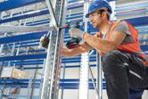 A person wearing safety equipment uses a cordless drill to assemble metal shelving.