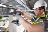 A person wearing safety equipment tightens bolts on a steel beam with a cordless drill.