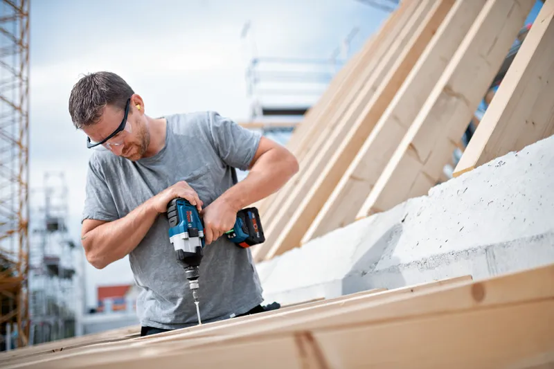 Person wearing safety equipment uses a cordless impact wrench to drive a screw into wood.