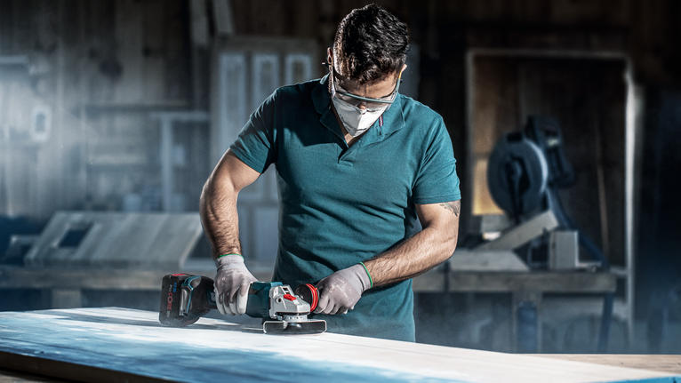 A person wearing safety equipment uses a cordless grinder on a wooden surface.
