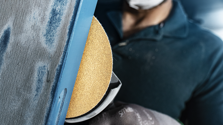 A person wearing safety equipment sands a metal surface with a rotary sander.