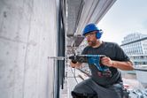 A person wearing safety equipment drills into concrete with a cordless rotary hammer.