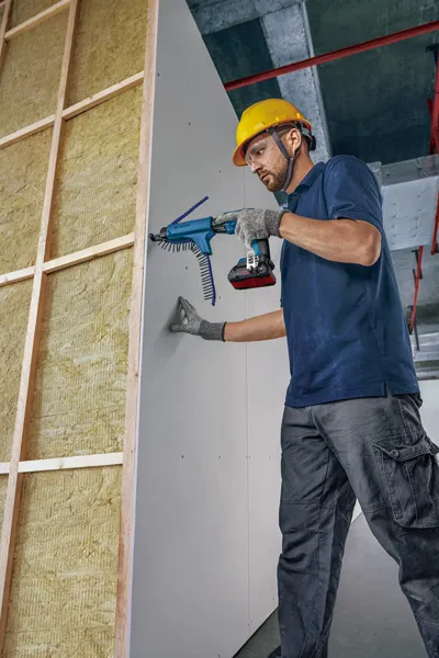 A person wearing safety equipment secures drywall to a frame using an automatic screw gun.