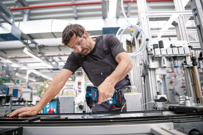 A person wearing safety equipment uses a cordless drill on an industrial assembly line.