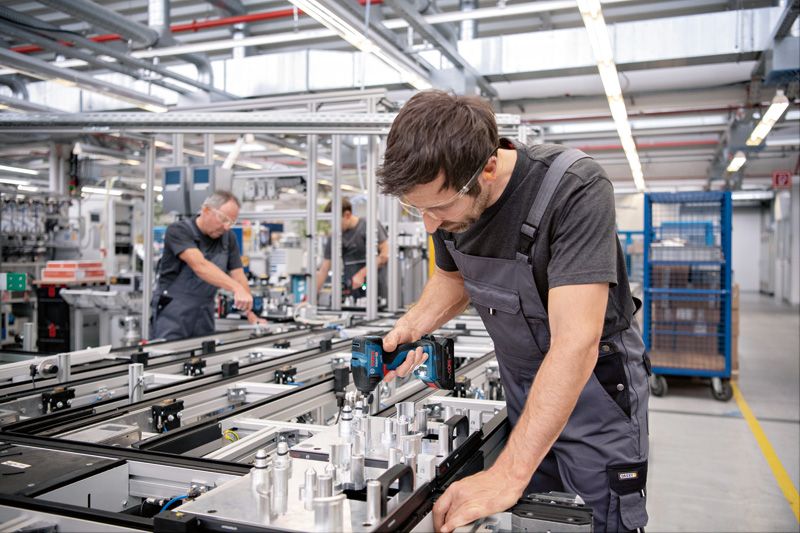 A person wearing safety equipment uses a cordless drill on an assembly line.