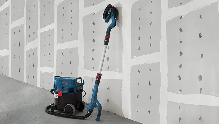 Drywall sander connected to a dust extractor resting against a finished wall.