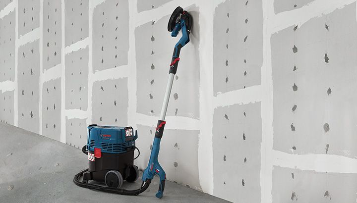 Drywall sander connected to a dust extractor resting against a finished wall.