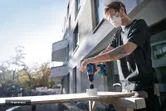 A person wearing safety equipment uses a power drill to cut a hole in a board.