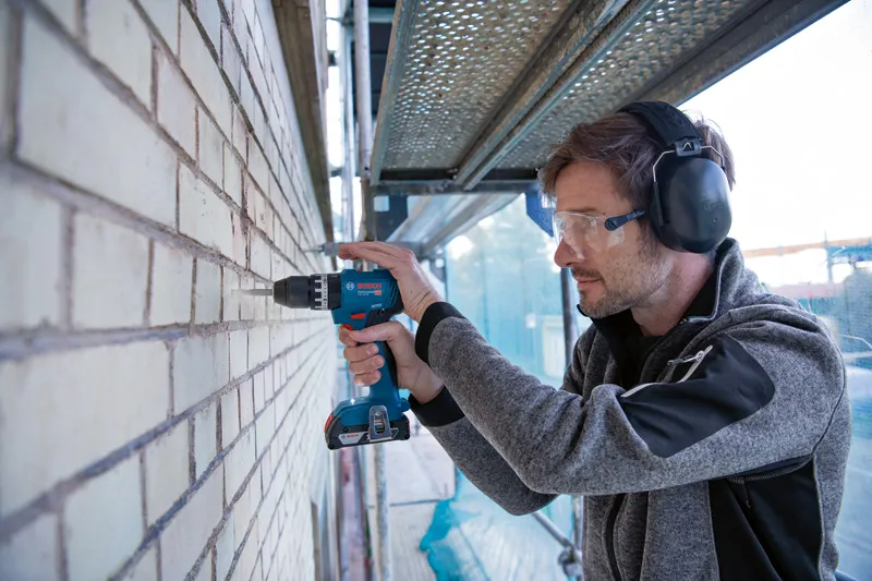 Person wearing safety equipment drills into a brick wall with a cordless power tool.