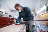 A person wearing safety equipment sands a wooden boat deck using a random orbit sander.