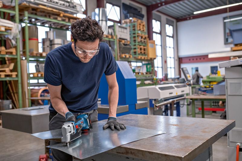 A person wearing safety equipment cuts sheet metal with a cordless metal shear.