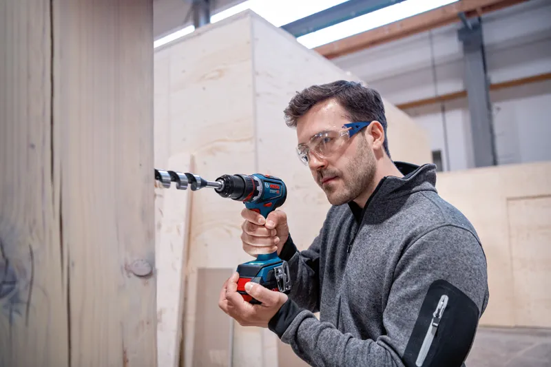 Person wearing safety equipment drills into wood using a cordless drill driver.