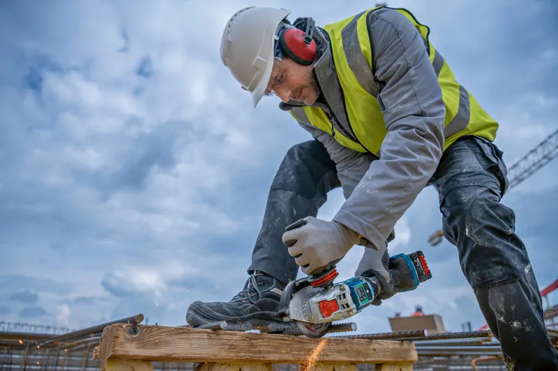 A person wearing safety equipment cuts rebar with a cordless angle grinder.