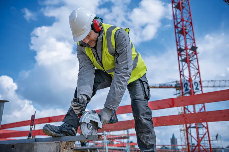 A person wearing safety equipment uses a cordless angle grinder to cut metal on a construction site.