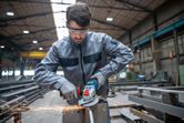A person wearing safety equipment grinds metal with a cordless angle grinder in a factory.
