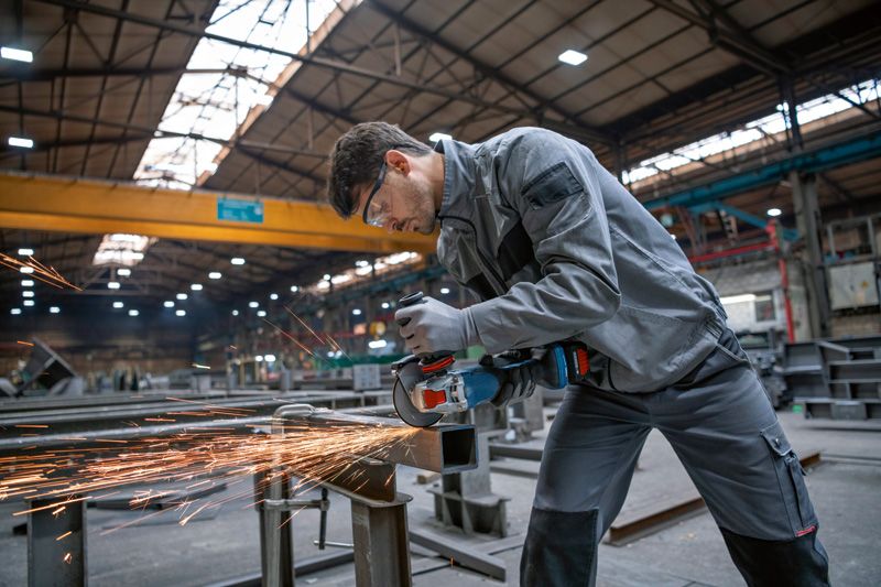 A person wearing safety equipment grinds metal, creating sparks in a large workshop.