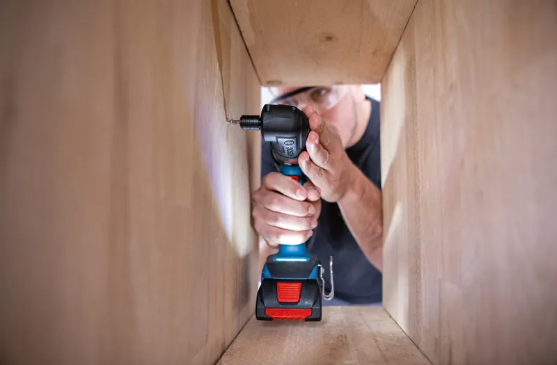 A person wearing safety equipment uses a cordless drill to drive a screw inside a wooden frame.
