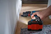 A person drills into a baseboard using a cordless drill on a wooden floor.