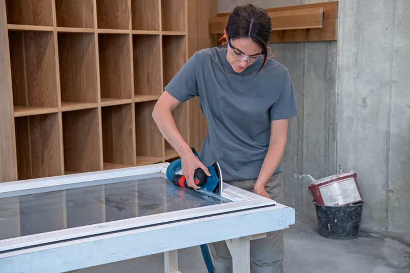 A person wearing safety equipment sands a window frame with a cordless orbital sander.