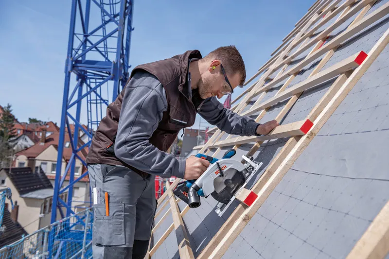 A person wearing safety equipment uses a circular saw to cut wood on a sloped roof.