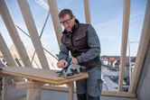 A person wearing safety equipment cuts a wooden board with a circular saw on a construction site.