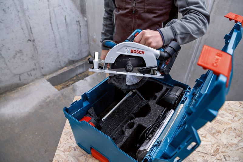 A person places a cordless circular saw into a blue tool case on a workbench.
