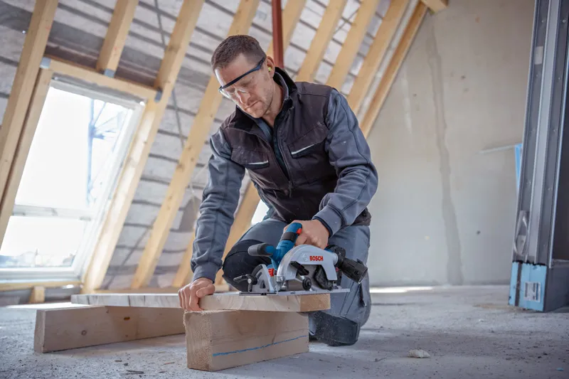 Person wearing safety equipment cuts wooden beam with a circular saw in an attic.